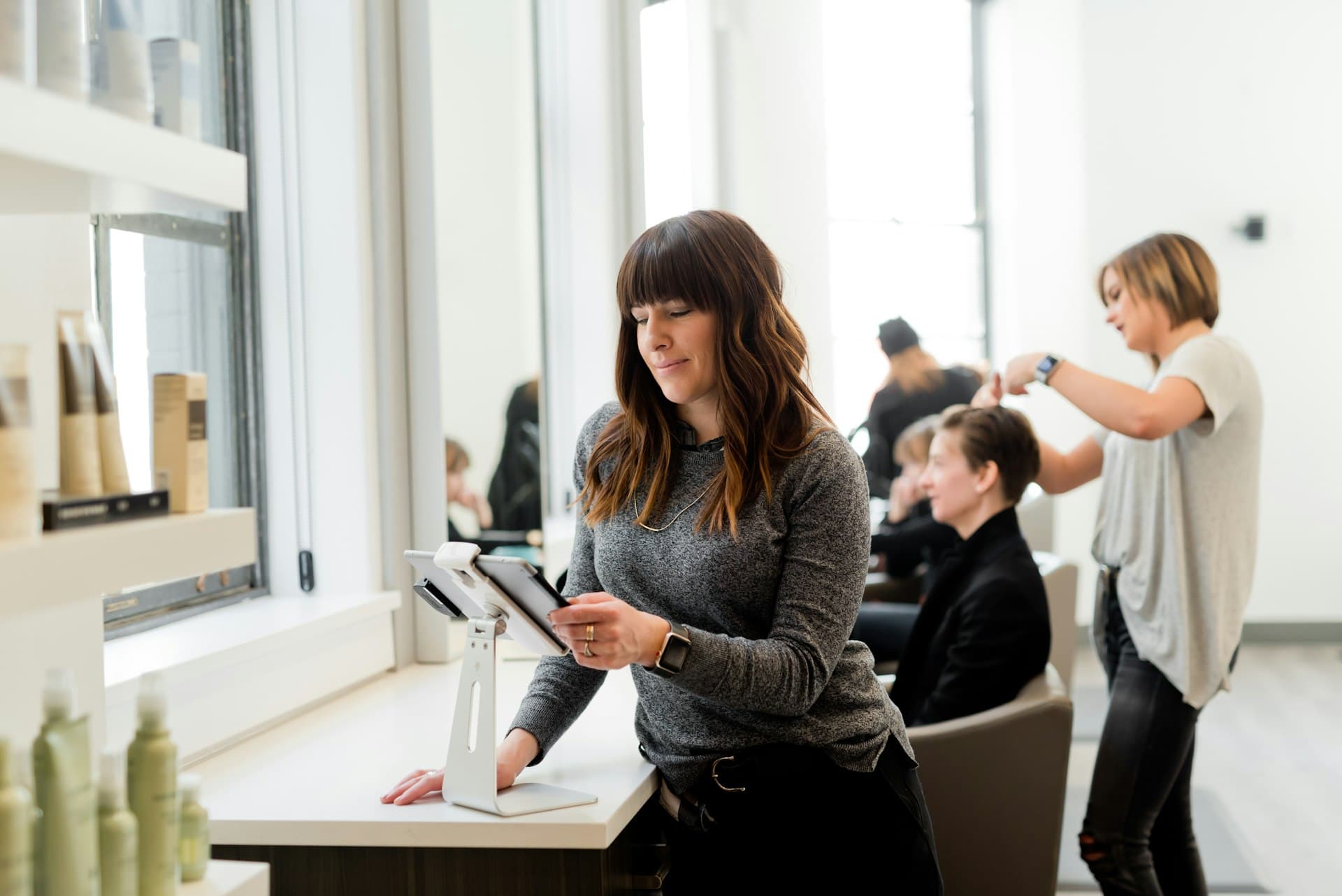 A salon owner checks a tablet at her front desk while her team works behind her